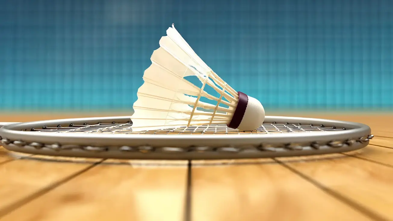 Close-up of a badminton shuttlecock resting on the strings of a racket on a wooden court floor, shot from low angle with the net blurred in the background