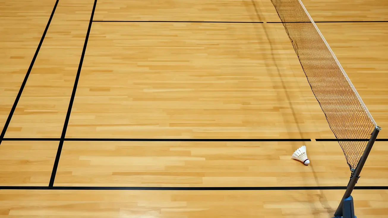 Overhead view of an empty badminton court showing the full court lines, net, and a shuttlecock resting near the service line on a wooden indoor surface