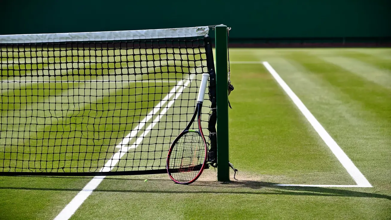 A tennis racket resting against the net post on an empty outdoor grass court, morning light casting long shadows across the surface